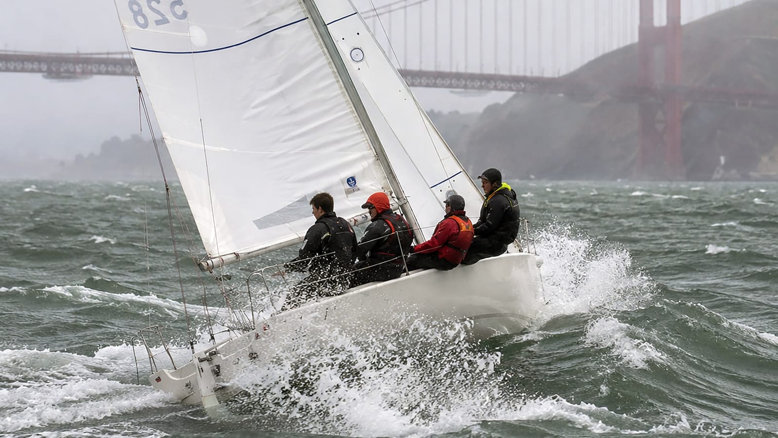 A J/80 heeling hard with its crew hiking out, the Golden Gate Bridge in the mist behind