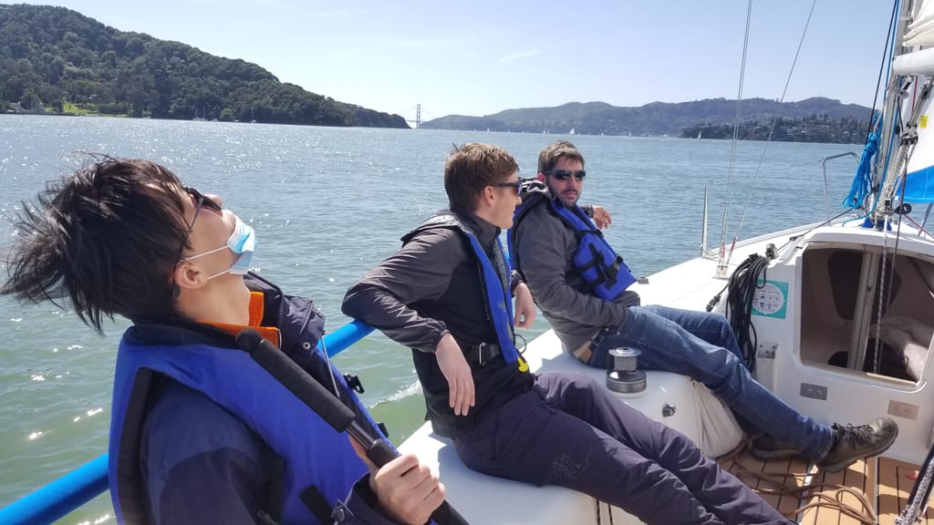 Three sailors on the rail of a keelboat with the Golden Gate Bridge in the distance